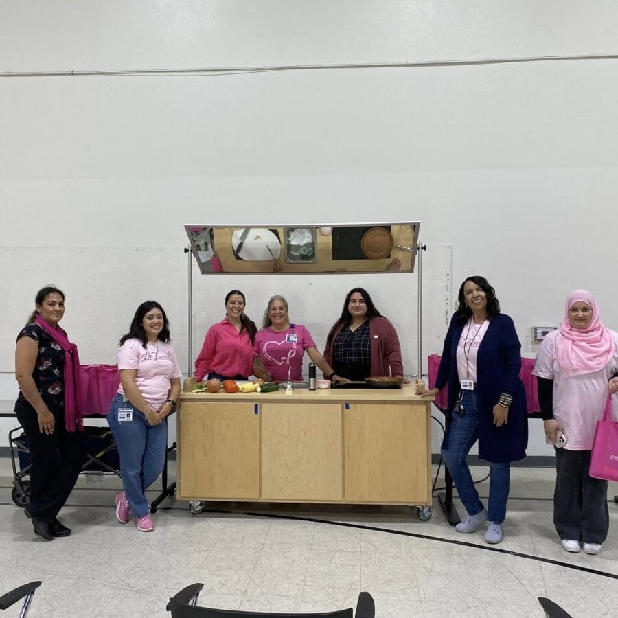Seven women stand in a row in front of a kitchen demonstration setup in a gymnasium, wearing pink or holding pink bags.
